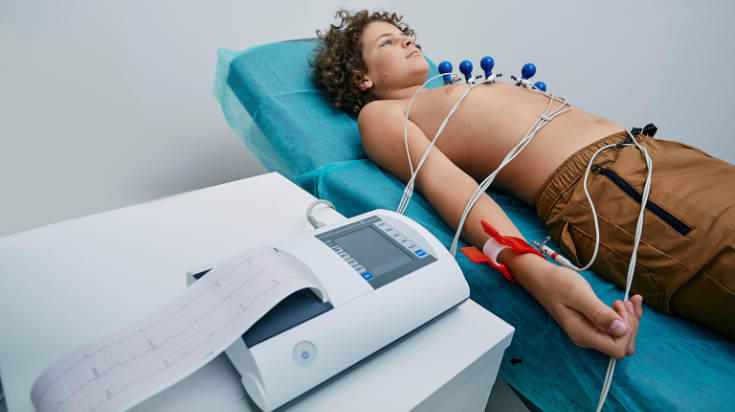 A patient lies on an examination table connected to ECG electrodes during a heart monitoring test.