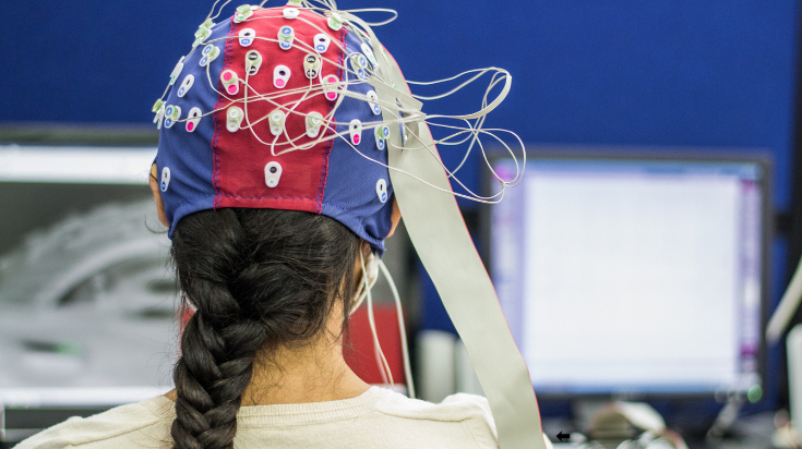 A person wearing an EEG cap with multiple electrodes sits in front of computer monitors during a brain activity recording session.