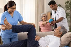 A caregiver assists an elderly man with leg exercises while another healthcare worker helps an elderly woman in the background.